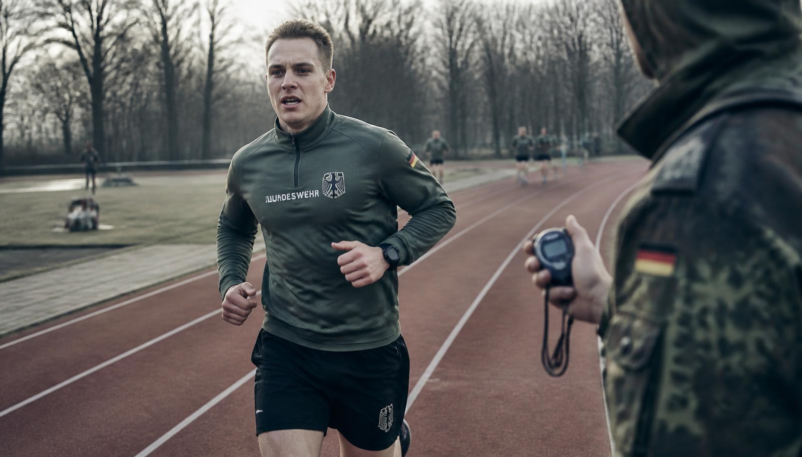 Soldat läuft den 1.000-Meter-Lauf des Basis-Fitness-Tests auf einer Tartanbahn, ein Ausbilder nimmt die Zeit