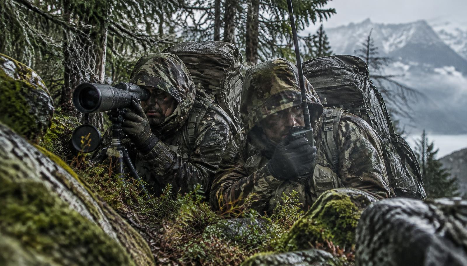Zwei Soldaten in Alpentarn in einem getarnten Beobachtungsposten im nassen Bergwald, mit Spektiv und Funkgerät.